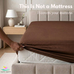 A person's hand pulls a brown fitted sheet over a white mattress on a bed with a beige upholstered headboard. Text at the top reads 'This Is Not a Mattress It covers your mattress' and a Happy Matty logo appears in the bottom left corner.