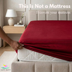 A fitted mattress cover in deep red is being pulled taut over a white mattress in a bedroom setting. White text at the top reads 'This Is Not a Mattress It covers your mattress', and the Happy Matty logo appears in the bottom-left corner.