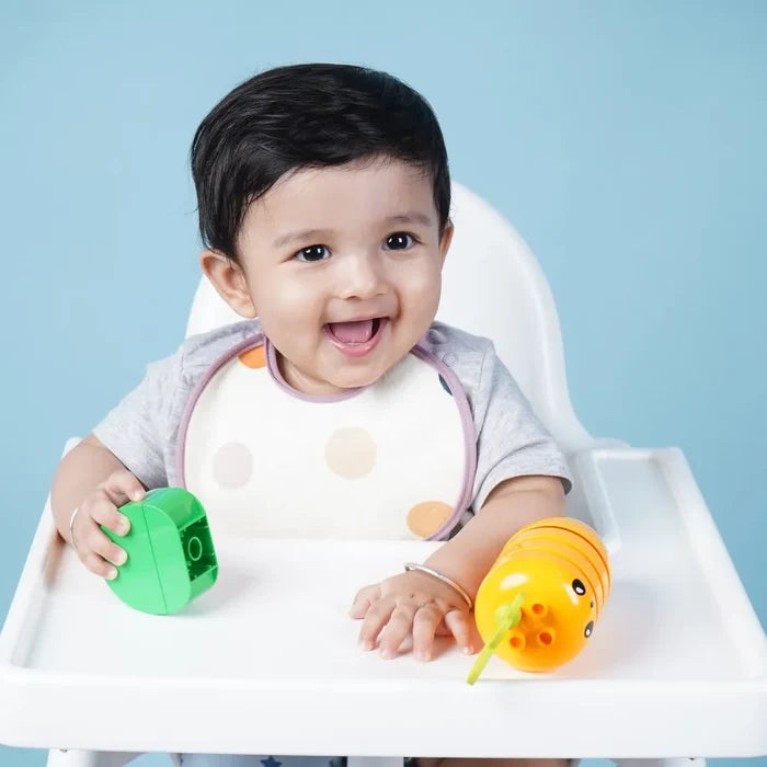 Smiling baby sitting in a high chair holding colourful toys while wearing a feeding bib