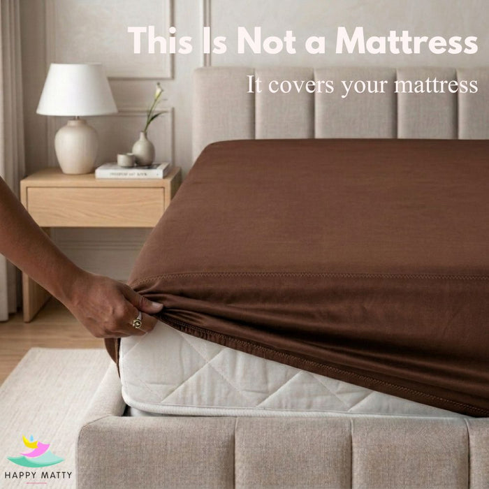 A person's hand pulls a brown fitted sheet over a white mattress on a bed with a beige upholstered headboard. Text at the top reads 'This Is Not a Mattress It covers your mattress' and a Happy Matty logo appears in the bottom left corner.